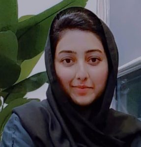 Young woman wearing a black hijab, smiling softly at the camera with green plant leaves in the background.